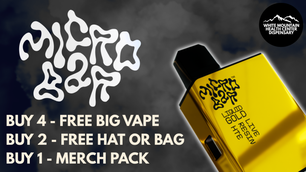 A promotional marketing graphic featuring a dark, smoky backdrop with stylized white typography spelling "MICROBAR". The right side displays a metallic gold Microbar "Liquid Gold Live Resin HTE" disposable vape device alongside the circular White Mountain Health Center Dispensary logo. Text on the left outlines tiered promotional offers: "BUY 4 - FREE BIG VAPE", "BUY 2 - FREE HAT OR BAG", and "BUY 1 - MERCH PACK".